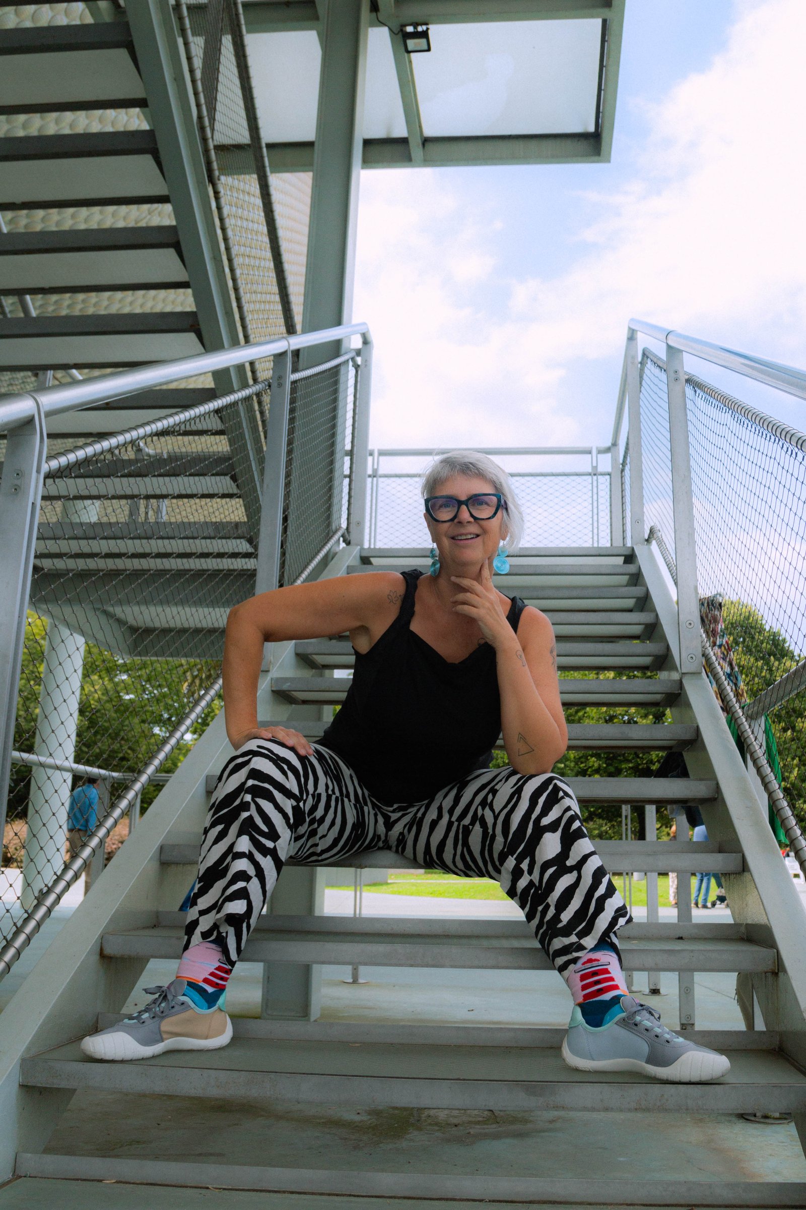 La fundadora de GELB sentada en una escalera metálica exterior, en una pose tranquila y reflexiva, con una mano cerca de la barbilla.