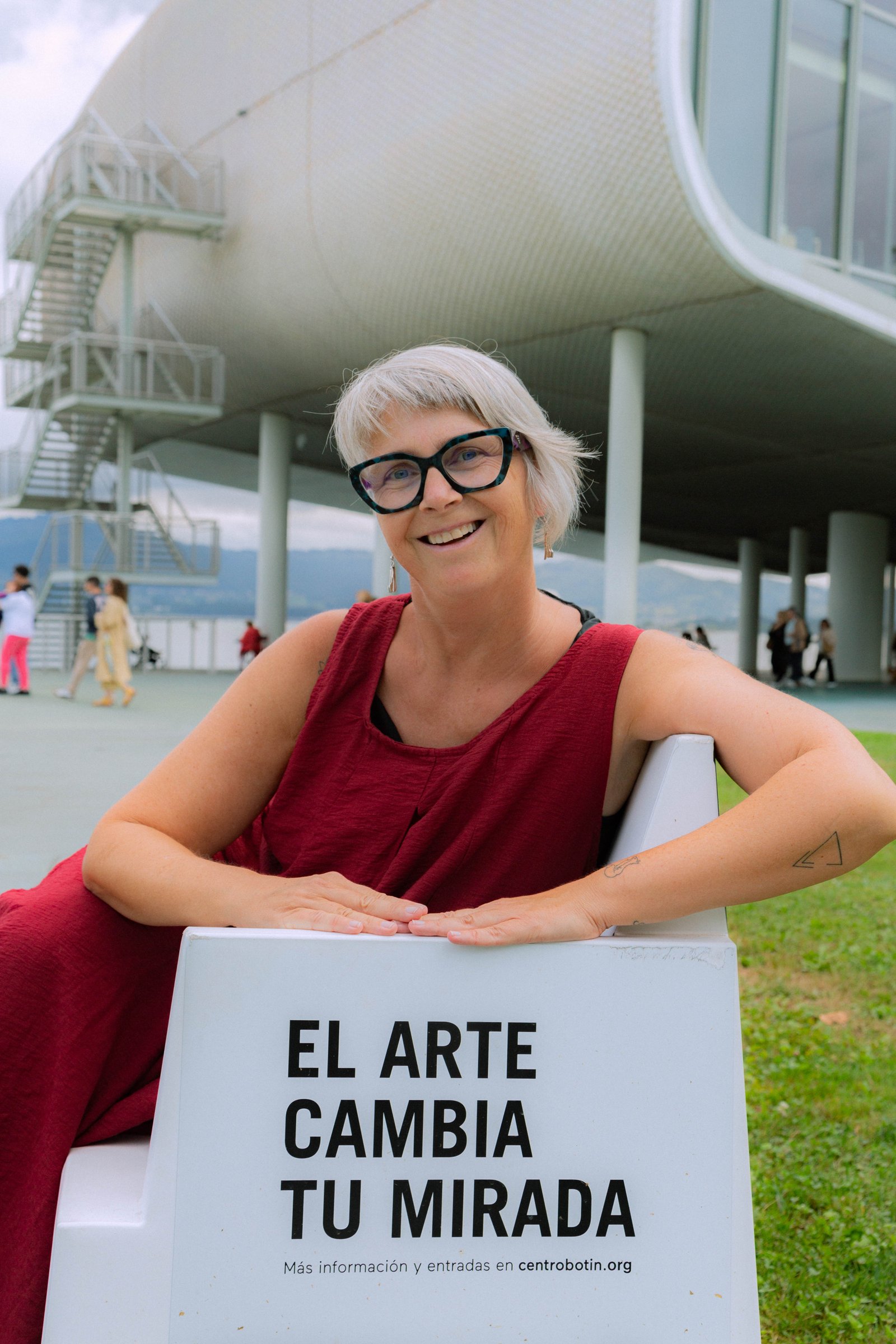 La fundadora de GELB apoyada en un cartel junto a un edificio de líneas curvas, sonriendo al aire libre.